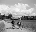 Två barn, en pojke och en flicka cyklar på en grusväg. Pojken har en resväska på pakethållaren, flickan har cykelväska. I bakgrunden ett hus med vita knutar och en lada.