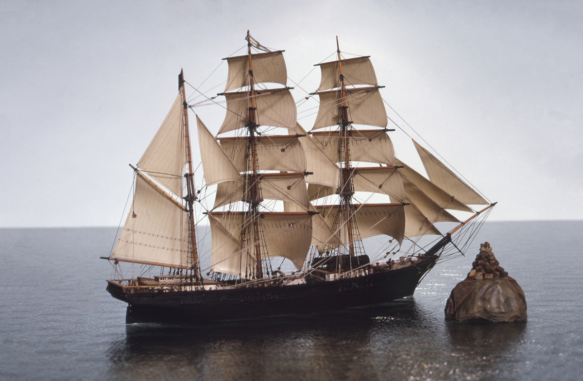 Fartygsmodell, vattenlinjemodell, hel, i block av trä, tremastade barkskeppet Hudiksvall av Hudiksvall (1864). Svartmålat skrov med galjonsbild föreställande kvinnohuvud, riggad med stående och löpande rigg, segel av papper. Två vita däckshus, två svarta livbåtar, namnvimpel på stortoppen, svensk flagga på mesantoppen.