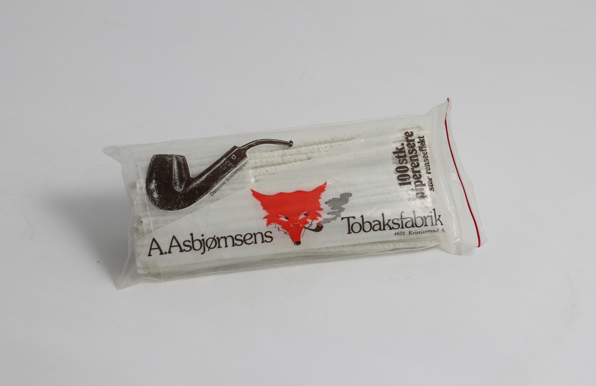Gjennomsiktig plastpose med piperensere. På siden er det et bilde av en pipe og et revehode, sammen med skriften "100 stk Piperensere" og "A. Asbjørnsens tobaksfabrik"