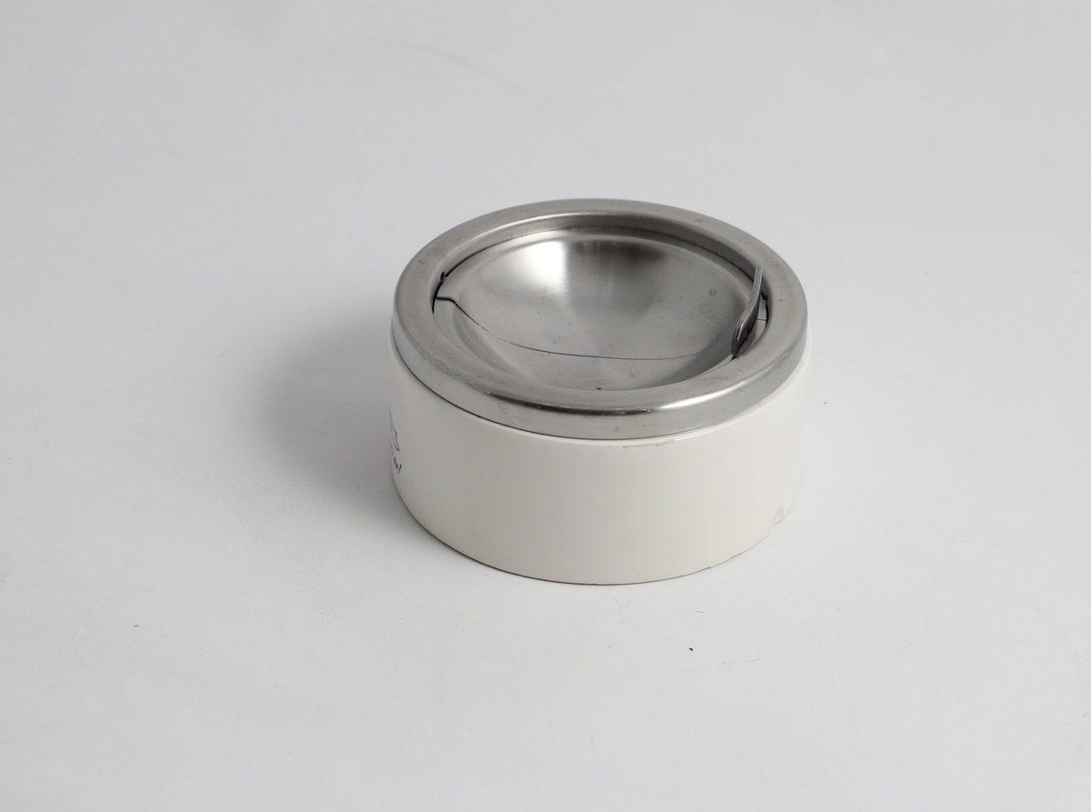 SIrkulært hvitt askebeger av plast med lokk av metall. Lokket har åpnemekanisme som slipped aske ned i begeret. På siden av plastbegeret er det bilde av en rev over tekst.