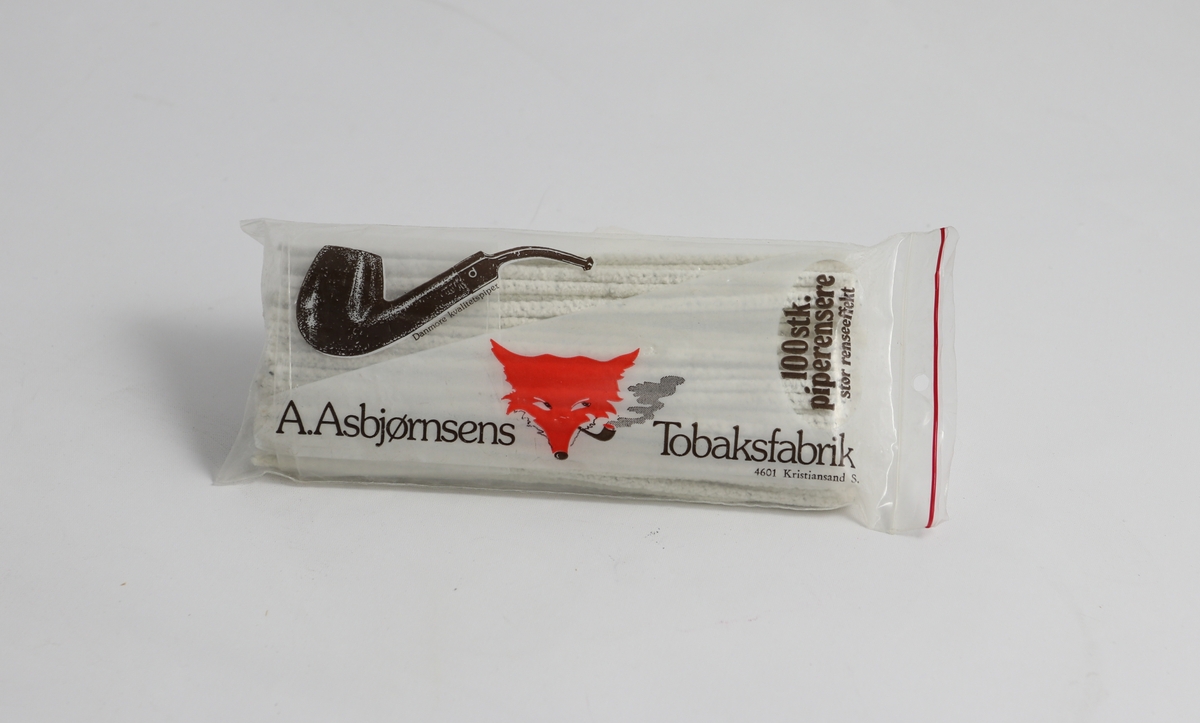 Gjennomsiktig plastpose med piperensere. På siden er det et bilde av en pipe og et revehode, sammen med skriften "100 stk Piperensere" og "A. Asbjørnsens tobaksfabrik"