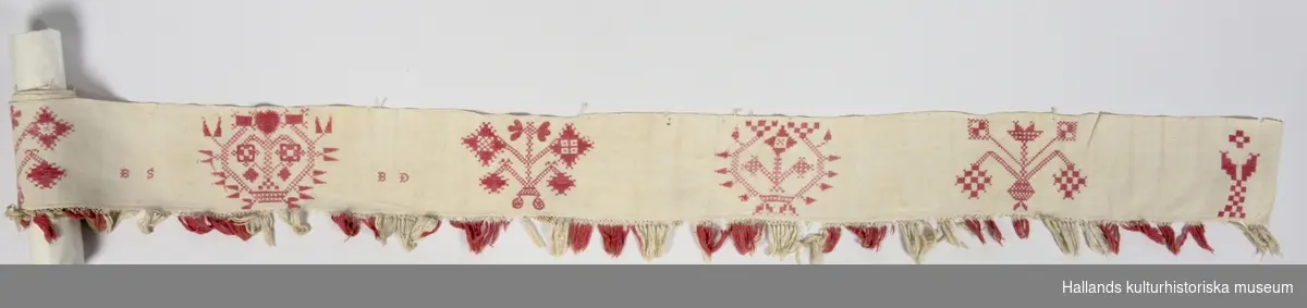 Hallandssöm. Mönster, mestadels i korsstygn broderat i rött. Fransar i vitt och rött. Hängklädet är blekt och slitet. Märkning: "BS BD" står för Britta Stina Börjesdotter. Bredd med frans: 155 mm. Hör ihop med föremål VMF043988