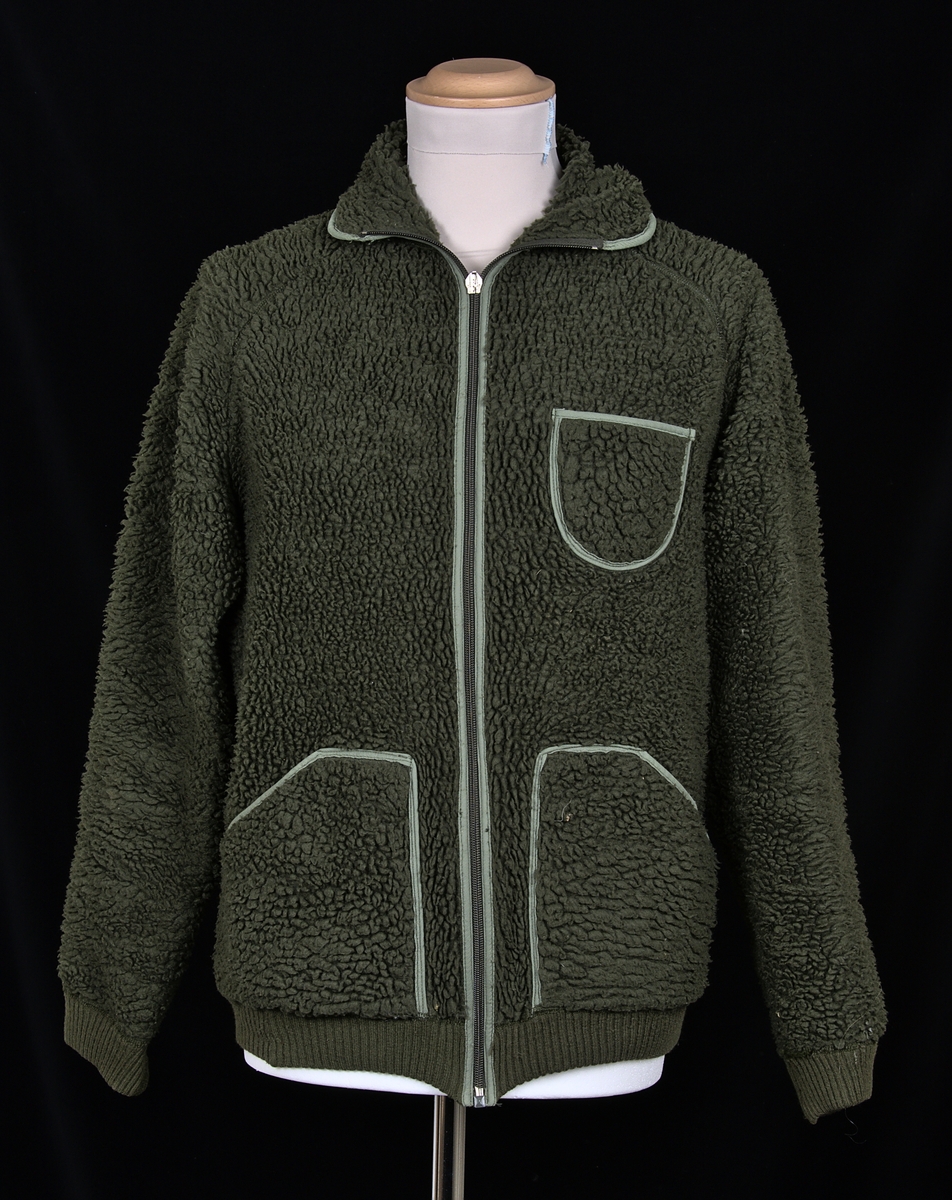 Jacka i mörkgrön fleece av polyester/akryl. Tjock lugg på utsidan. Två höftfickor, samt en bröstficka framtill. Stängs med hjälp av dragkedja.