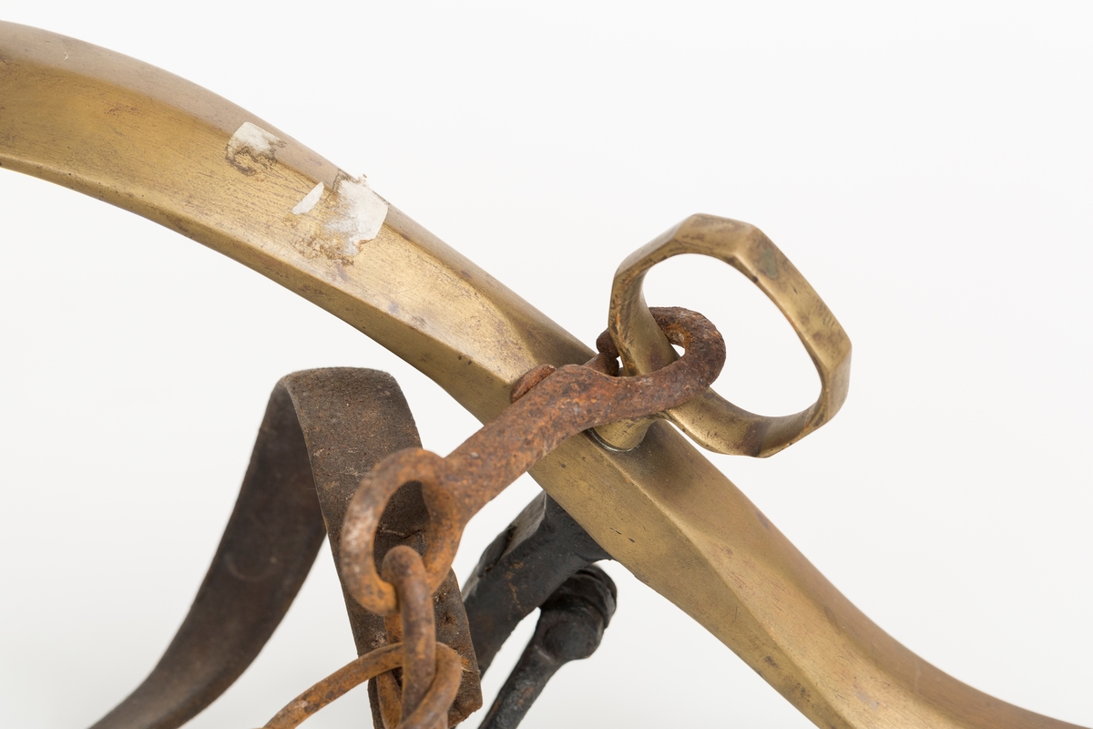 Høvre i smidd stål trukket med messing med påloddete messingringer/-fester. Stykke av lærrem festet med D-ring, lenkeledd og karabinkrok til messingring på høvret. Rund messingplate med bøyle er tredd inn på reimen.