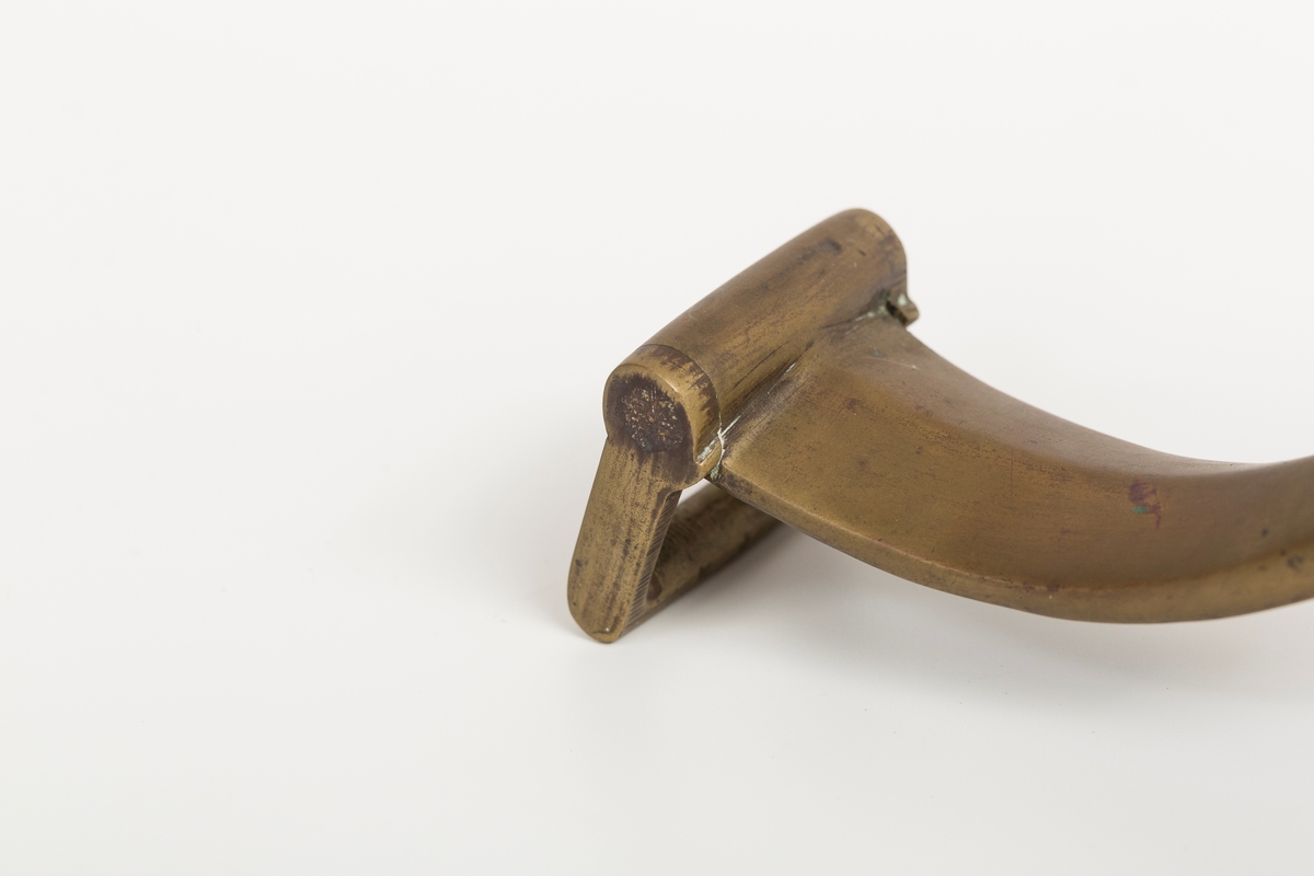 Høvre i smidd stål trukket med messing med påloddete messingringer/-fester. Stykke av lærrem festet med D-ring, lenkeledd og karabinkrok til messingring på høvret. Rund messingplate med bøyle er tredd inn på reimen.