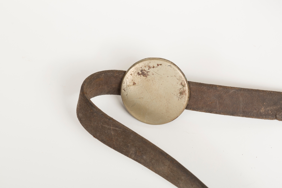 Høvre i smidd stål trukket med messing med påloddete messingringer/-fester. Stykke av lærrem festet med D-ring, lenkeledd og karabinkrok til messingring på høvret. Rund messingplate med bøyle er tredd inn på reimen.