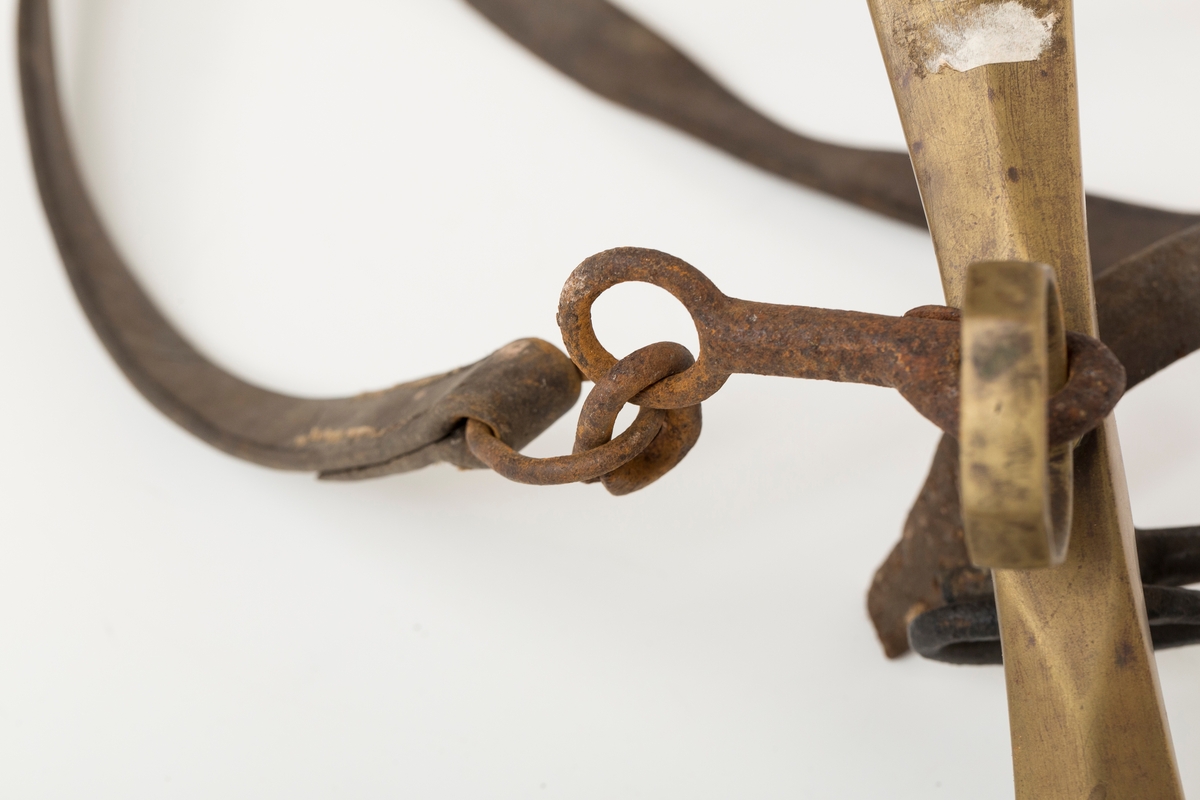Høvre i smidd stål trukket med messing med påloddete messingringer/-fester. Stykke av lærrem festet med D-ring, lenkeledd og karabinkrok til messingring på høvret. Rund messingplate med bøyle er tredd inn på reimen.