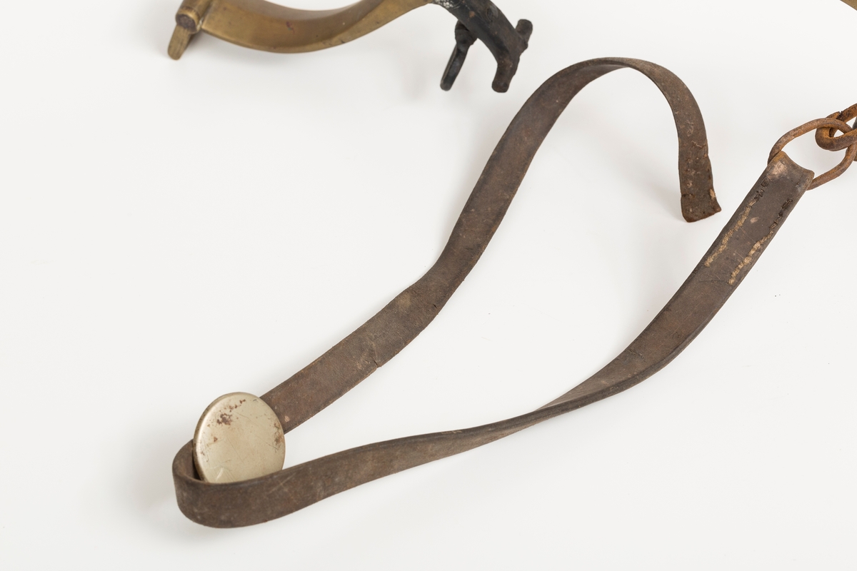 Høvre i smidd stål trukket med messing med påloddete messingringer/-fester. Stykke av lærrem festet med D-ring, lenkeledd og karabinkrok til messingring på høvret. Rund messingplate med bøyle er tredd inn på reimen.
