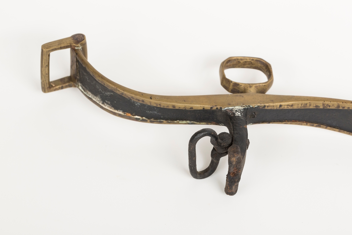 Høvre i smidd stål trukket med messing med påloddete messingringer/-fester. Stykke av lærrem festet med D-ring, lenkeledd og karabinkrok til messingring på høvret. Rund messingplate med bøyle er tredd inn på reimen.