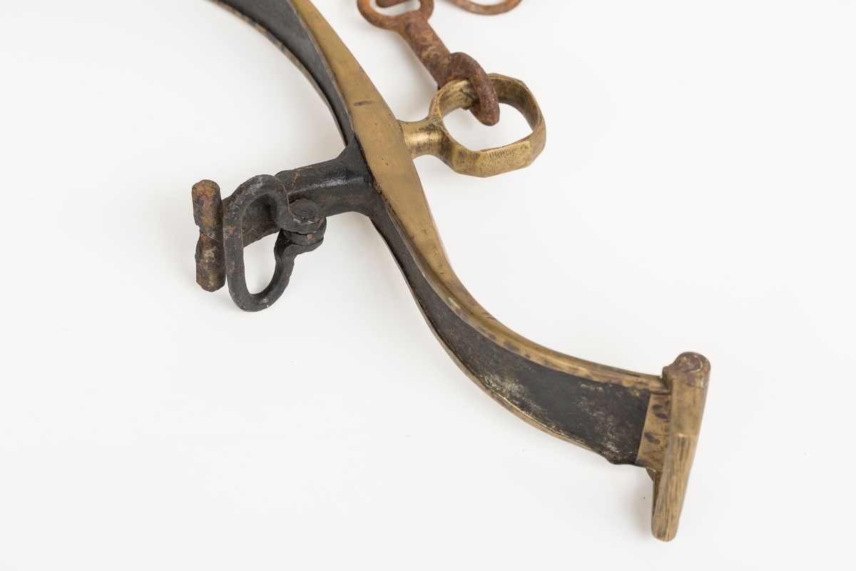 Høvre i smidd stål trukket med messing med påloddete messingringer/-fester. Stykke av lærrem festet med D-ring, lenkeledd og karabinkrok til messingring på høvret. Rund messingplate med bøyle er tredd inn på reimen.
