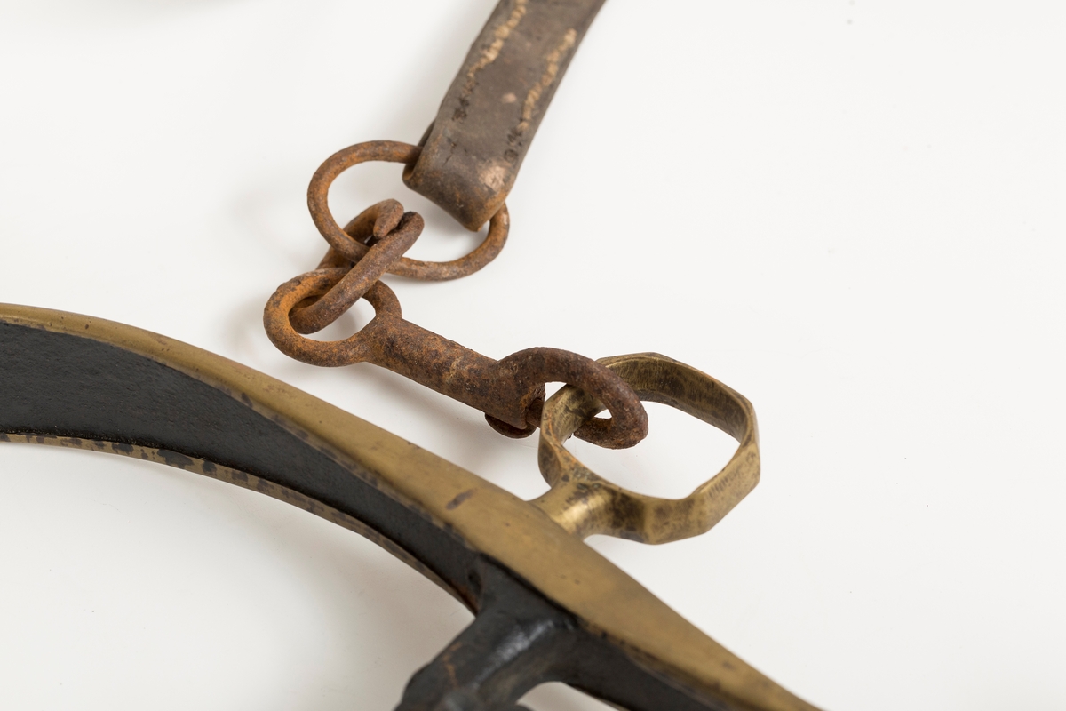 Høvre i smidd stål trukket med messing med påloddete messingringer/-fester. Stykke av lærrem festet med D-ring, lenkeledd og karabinkrok til messingring på høvret. Rund messingplate med bøyle er tredd inn på reimen.