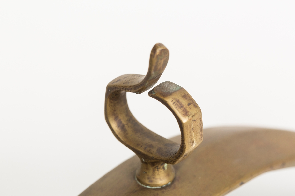 Høvre i smidd stål trukket med messing med påloddete messingringer/-fester. Stykke av lærrem festet med D-ring, lenkeledd og karabinkrok til messingring på høvret. Rund messingplate med bøyle er tredd inn på reimen.