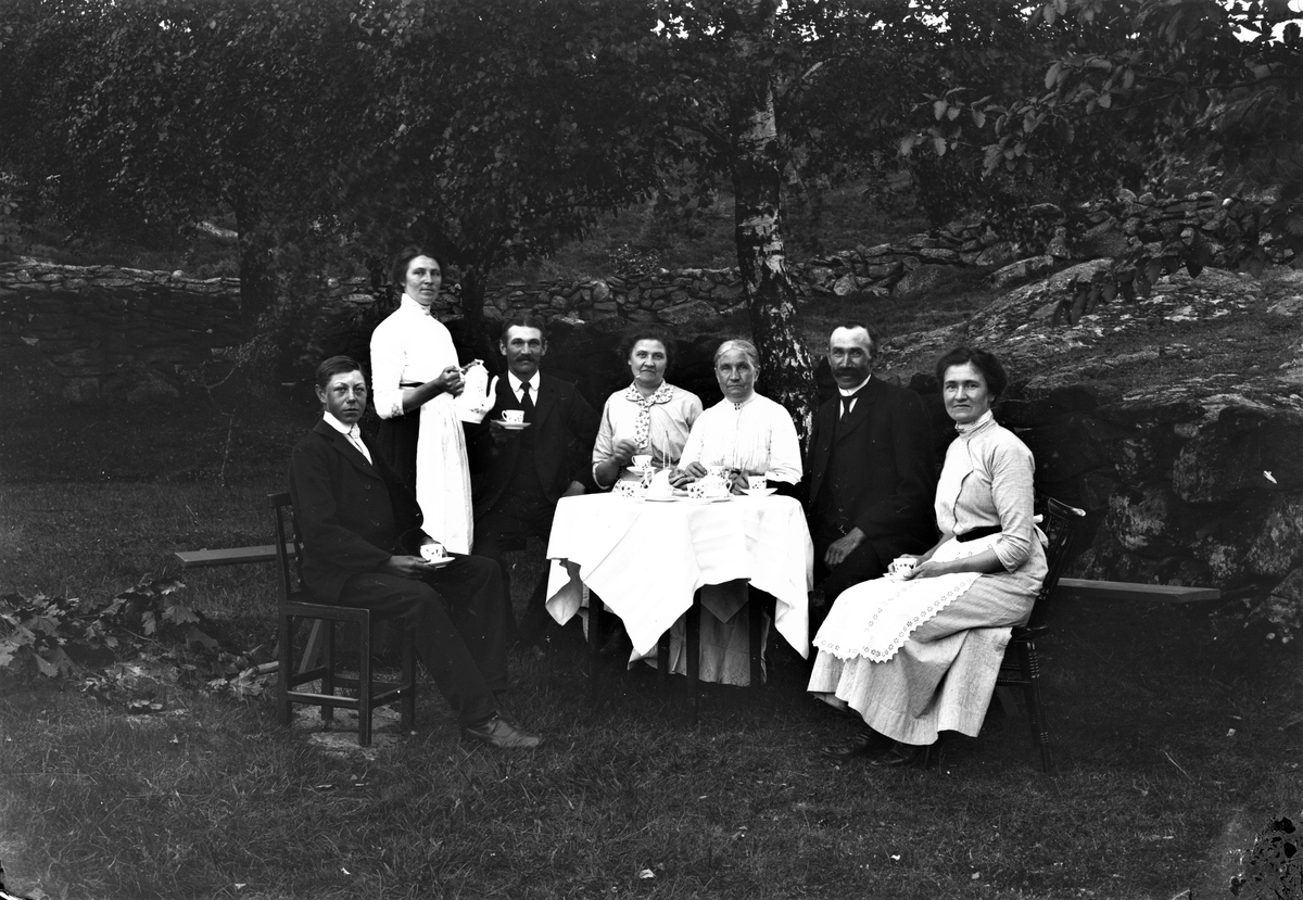 Folket på Kärskens i Grimmared har en kaffestund i trädgården. Julia håller i kaffekannan för att servera Axel och sedan följer Tilda, Josefin, Birger och Hulda. Ynglingen till vänster är dräng på gården. En stengärdsgård går upp längs slänten bakom dem och nedanför bänken ligger en hög med avklippta lönnkvistar.
(Se även bilderna GEA029, GEA112, GEA114 och GEA116)