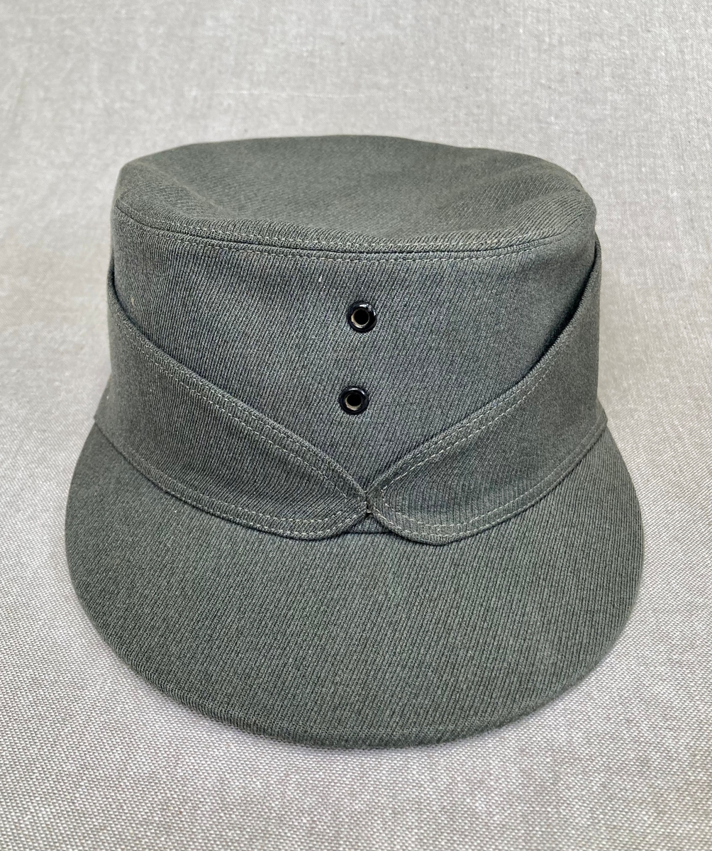 Grågrønn uniform med liggende krave og fire lommer, ridebukse og skilue (Finn Quales modell). Uniformen har bruksmerker, særlig på kraven. Luen er slitt, foret har løsnet. Alle gradstegn og knapper er fjernet fra jakken. God kvalitet på stoffet, muligens offisersuniform. (NB: Fremstår som grå på bildene, men har i virkeligheten en blass militærgrønn farge.)