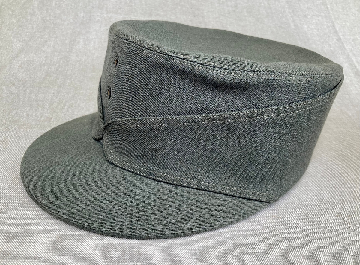 Grågrønn uniform med liggende krave og fire lommer, ridebukse og skilue (Finn Quales modell). Uniformen har bruksmerker, særlig på kraven. Luen er slitt, foret har løsnet. Alle gradstegn og knapper er fjernet fra jakken. God kvalitet på stoffet, muligens offisersuniform. (NB: Fremstår som grå på bildene, men har i virkeligheten en blass militærgrønn farge.)