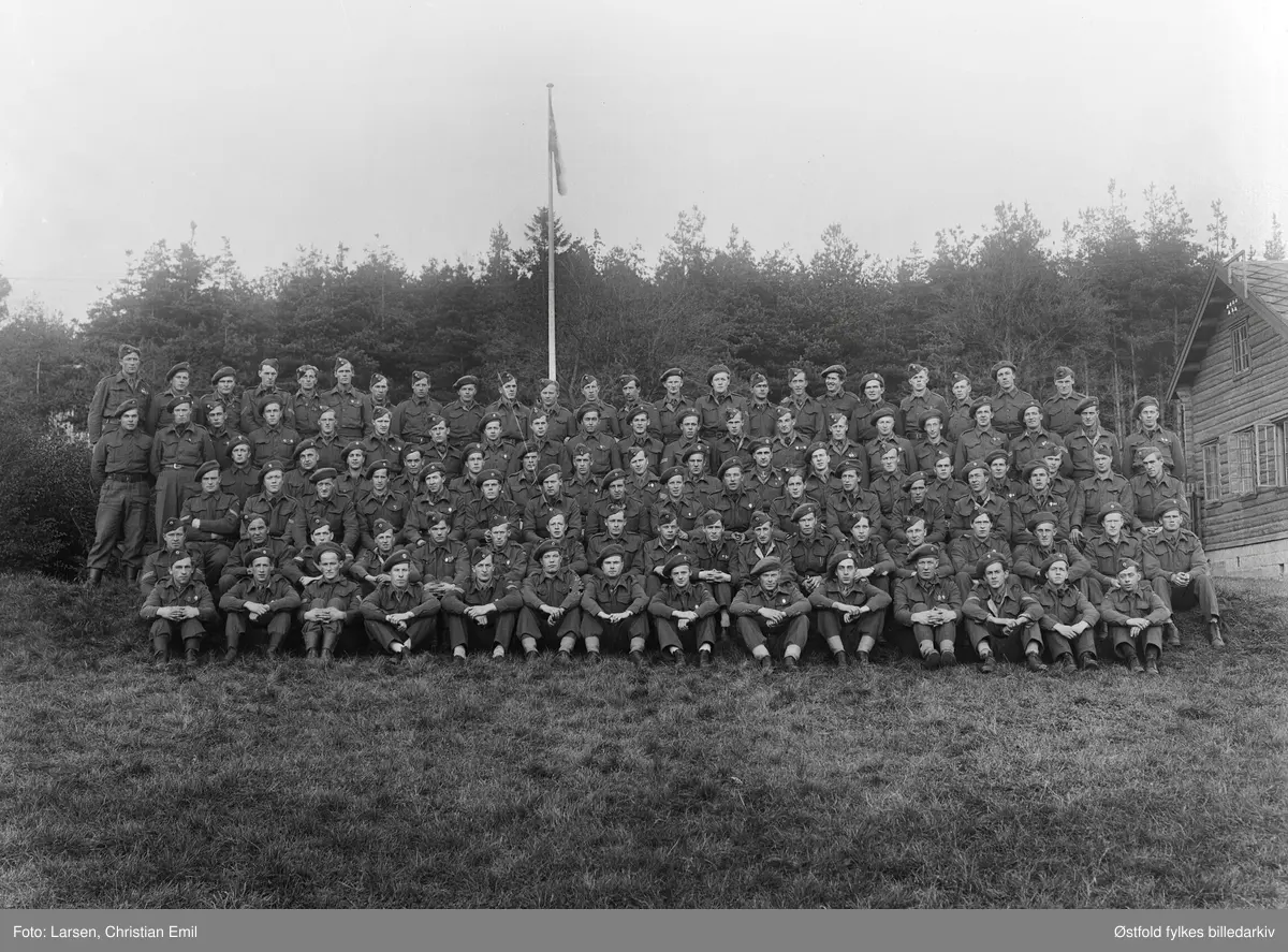 Tuneleiren i Tune 1945. Stort gruppebilde av soldater i uniform. Tuneleiren var tyskernes brakkeleir som lå på grunnen til Tune prestegård i Tune.
Man kan skimte «offiserboligen» i skogen helt til høyre på bildet, Tuneveien 46.