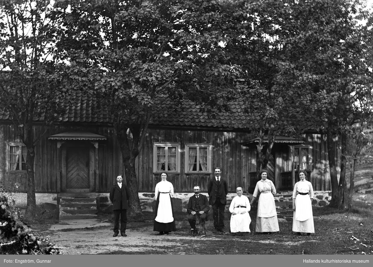 Folket på Kärskens i Grimmared står utanför manhuset och raden av lönnar som står tätt inpå huset.  Från höger ses Hulda, Tilda, Josefin, Axel, Birger, Julia och gårdsdrängen. De dubbla ingångarna antyder att byggnaden troligen är en s k loftgångsstuga, dvs en tidigare högloftsstuga där taket på ryggåsstugan i mitten har höjts upp i linje med de högre sidoloftens tak. Loften hade separata ingångar som ledde till smala hallar varifrån både stuga och loft nåddes och här har båda sparats efter ombyggnaden.
(Se även bilderna GEA029, GEA112, GEA114 och GEA115)