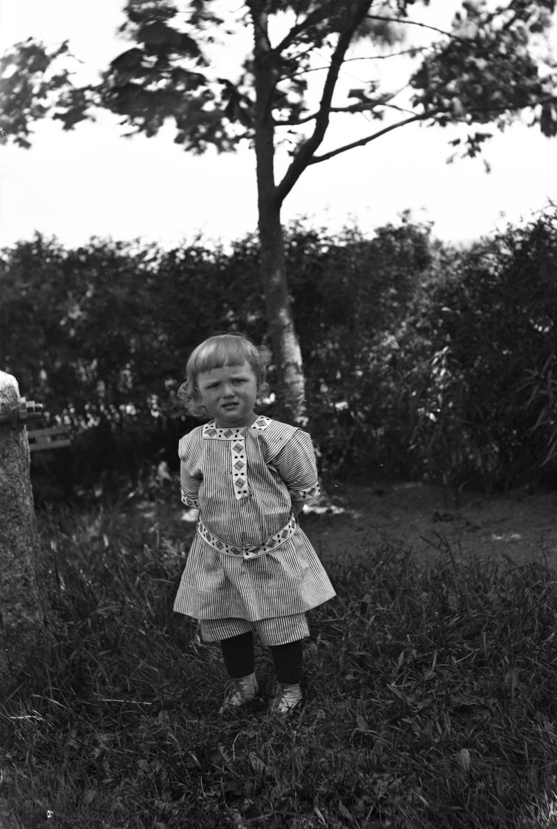 En liten pojke står i randig kolt med händerna på ryggen och undrande min mot fotografen. (Se även bildnr GEA135)