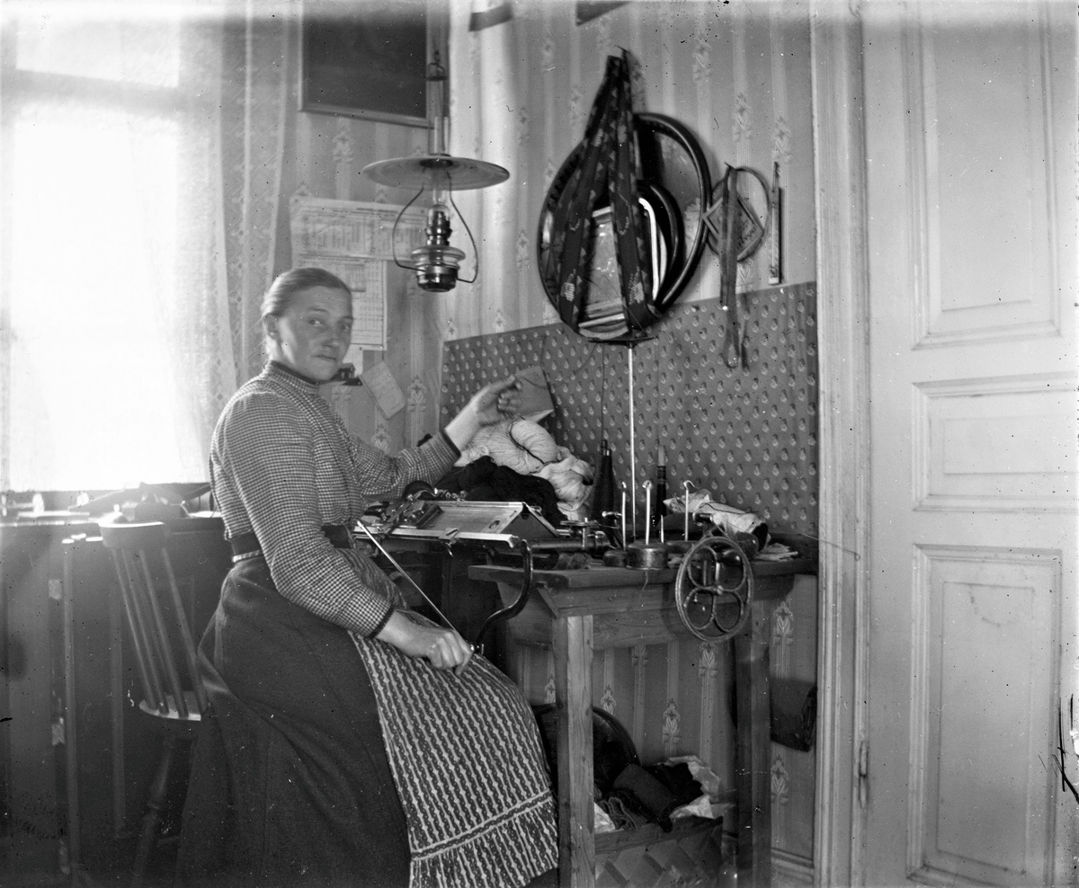 Gunnars mor Bertha vid stickmaskinen. Bertha har skyddat tapeten mot de skador stickningen kan åstadkomma och kvällstid har hon en fotogenlampa till belysning. Troligen ryms maskinen i köket eftersom det hänger brickor i brickband på väggen över den. 

I Viskadalen, och Sjuhäradsbygden i övrigt, förekom det s k förläggarsystemet. Förläggarna köpte levererade material och utrustning till textilarbeterskorna i hemmen runtom i trakten. Förläggarna samlade sedan in de färdiga produkterna.
