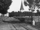Vy längs vägen genom Viby omkring 1905. Till vänster Källgården med ortens handelsbod, som vid tiden upplysningsvis drevs av bildens fotograf, Carl Sundström. Till höger församlingens kyrka som stått på platsen sedan 1100-talet.
