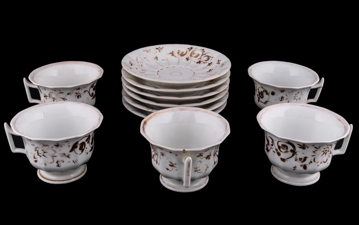 Kaffekoppar 5 st, med 6 st fat. Vitt gods med gulddekor av blommor och rocailler. Upptill utsvängd kopp, indragen mot foten. Uppsvängt öra, rakt avslutat nedåt. Uppsvängt fat.
