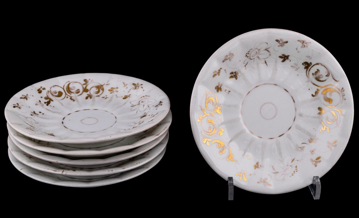 Kaffekoppar 5 st, med 6 st fat. Vitt gods med gulddekor av blommor och rocailler. Upptill utsvängd kopp, indragen mot foten. Uppsvängt öra, rakt avslutat nedåt. Uppsvängt fat.