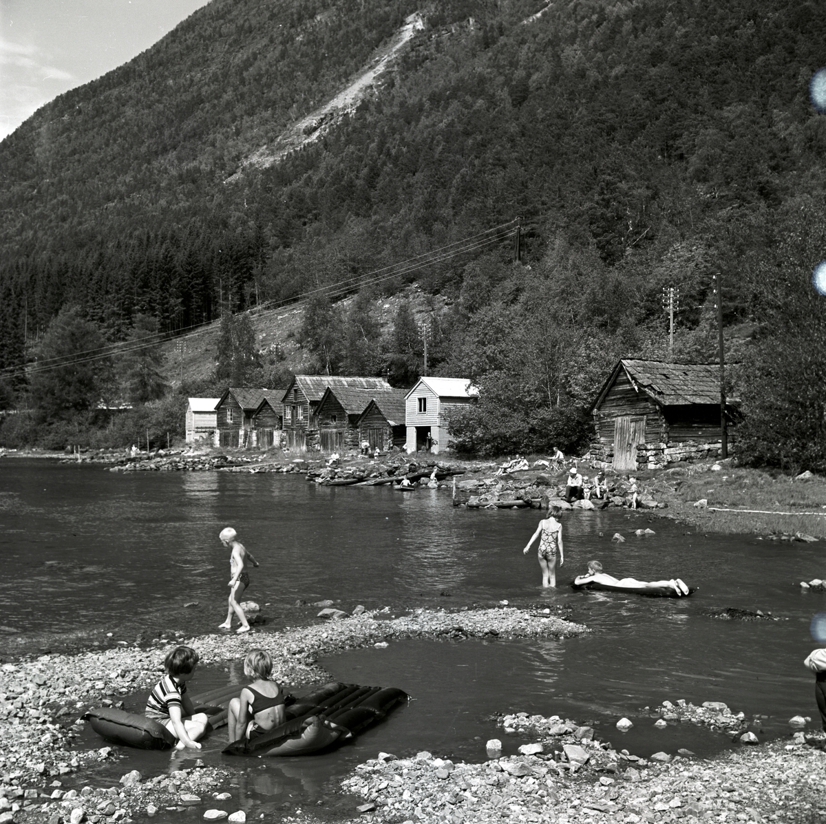 Foto av barn som bader i vannkanten. Langs stranda står eldre naust.
