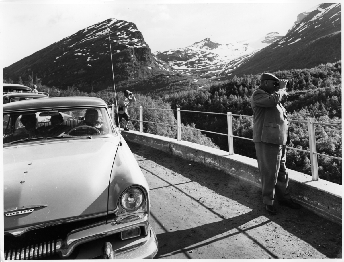 Foto av en mann som ser på utsikten ved Geiranger gjennom kikkert. En 1955 modell Plymouth står parkert ved veien.