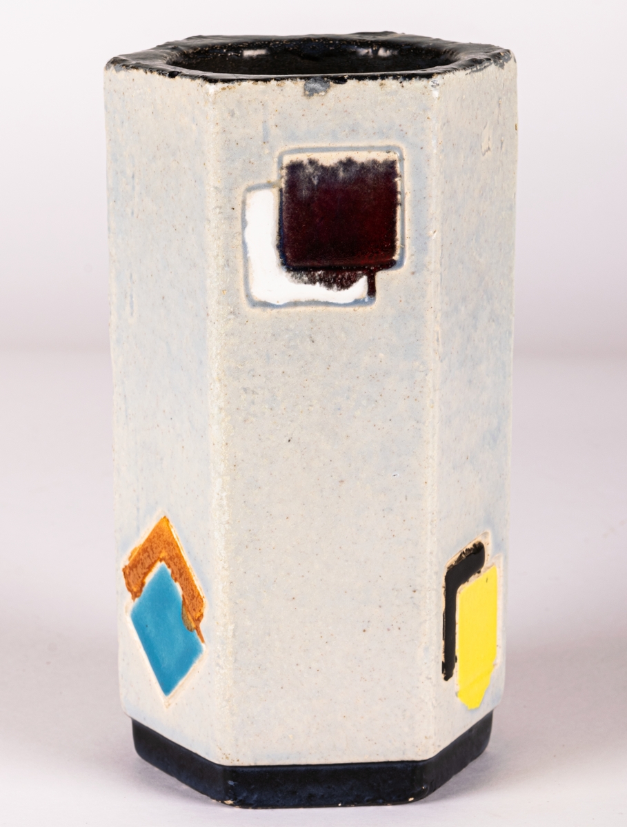 Sexkantig vas i chamottelera från Bo Fajans, modell 453, tillverkad 1953. Formgivare okänd men troligen Lillie Dahlgren, möjligen Tom Wilson. Ljus glasyr med dekor av fyrkantiga rutor i olika färger, svart sockeldel.
