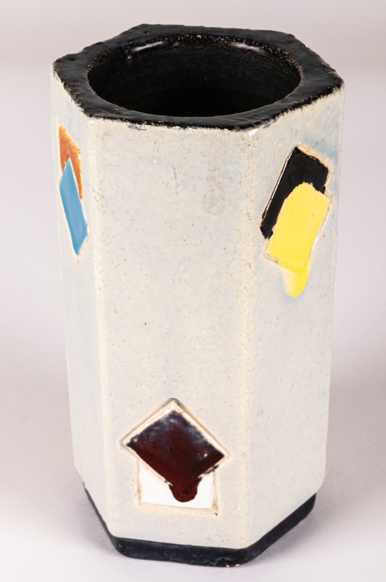 Sexkantig vas i chamottelera från Bo Fajans, modell 453, tillverkad 1953. Formgivare okänd men troligen Lillie Dahlgren, möjligen Tom Wilson. Ljus glasyr med dekor av fyrkantiga rutor i olika färger, svart sockeldel.