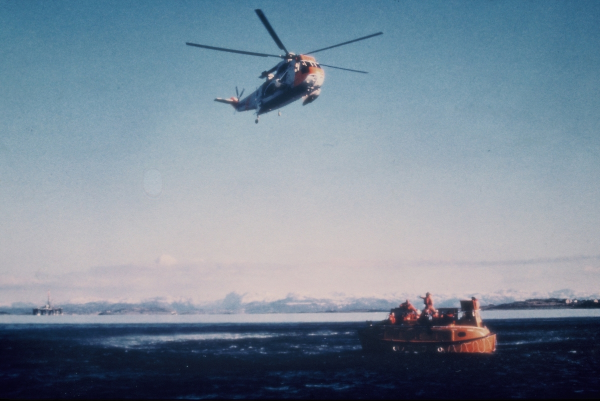 Redningsøvelse i en fjord på sørvestlandet. Et redningshelikopter sirkler over en livbåt med mannskap ombord. På bilde nr. 2 heises en livredder ned fra helikopteret.