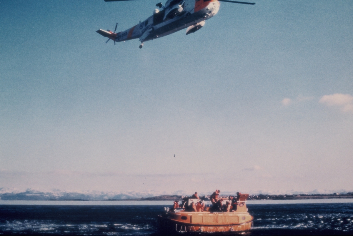 Redningsøvelse i en fjord på sørvestlandet. Et redningshelikopter sirkler over en livbåt med mannskap ombord. På bilde nr. 2 heises en livredder ned fra helikopteret.