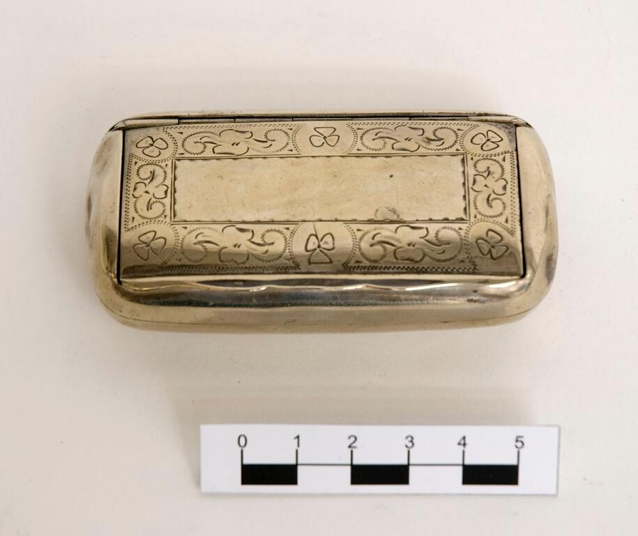 Snusdosa, av tennliknande material, rektangulär med konvexa sidor. Nedsänkt lock, med dekor av ristade treklöver, zigzaglinjer och blad. Gåva: Fru häradshövding Grufman, Stockholm 1960, (samling tillhört fadern distriktslantmätare A.H.L. Andersson, Sollefteå). 