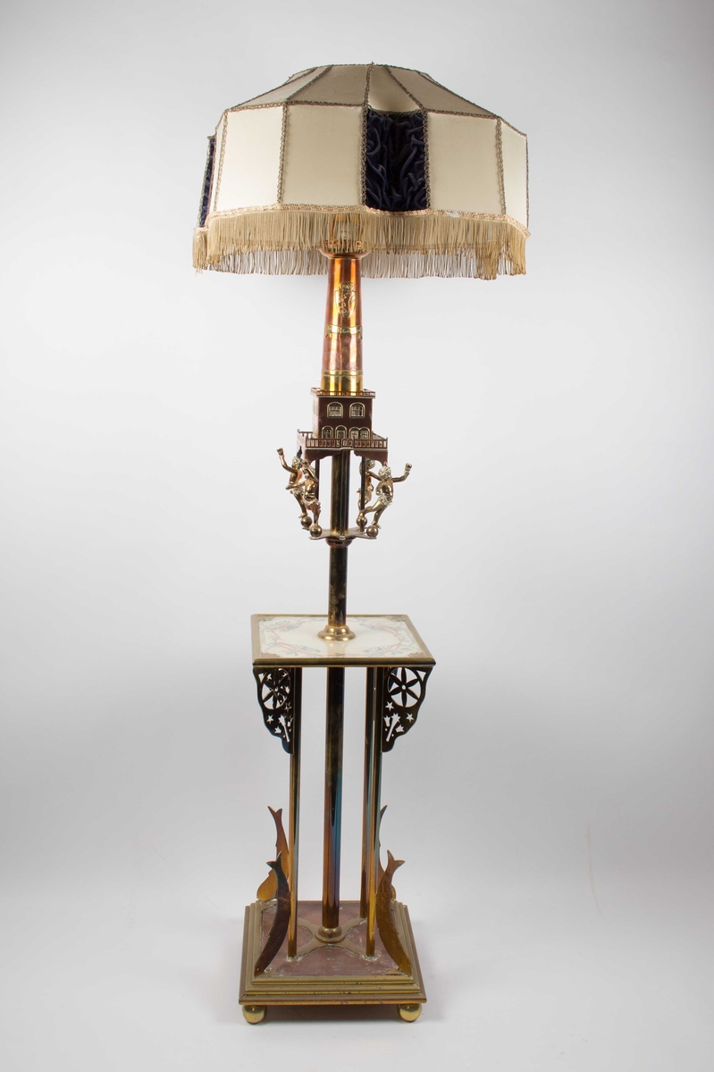 Stålampe i kobber og messing med tekstil lampeskjerm (uoriginal). Lampefoten er laget som et fyrtårn holdt oppe av noen små guttefigurer. I tillegg er det et lite bord med en bordduk i tekstil med motiv av fire fugler. Foten er i messing.