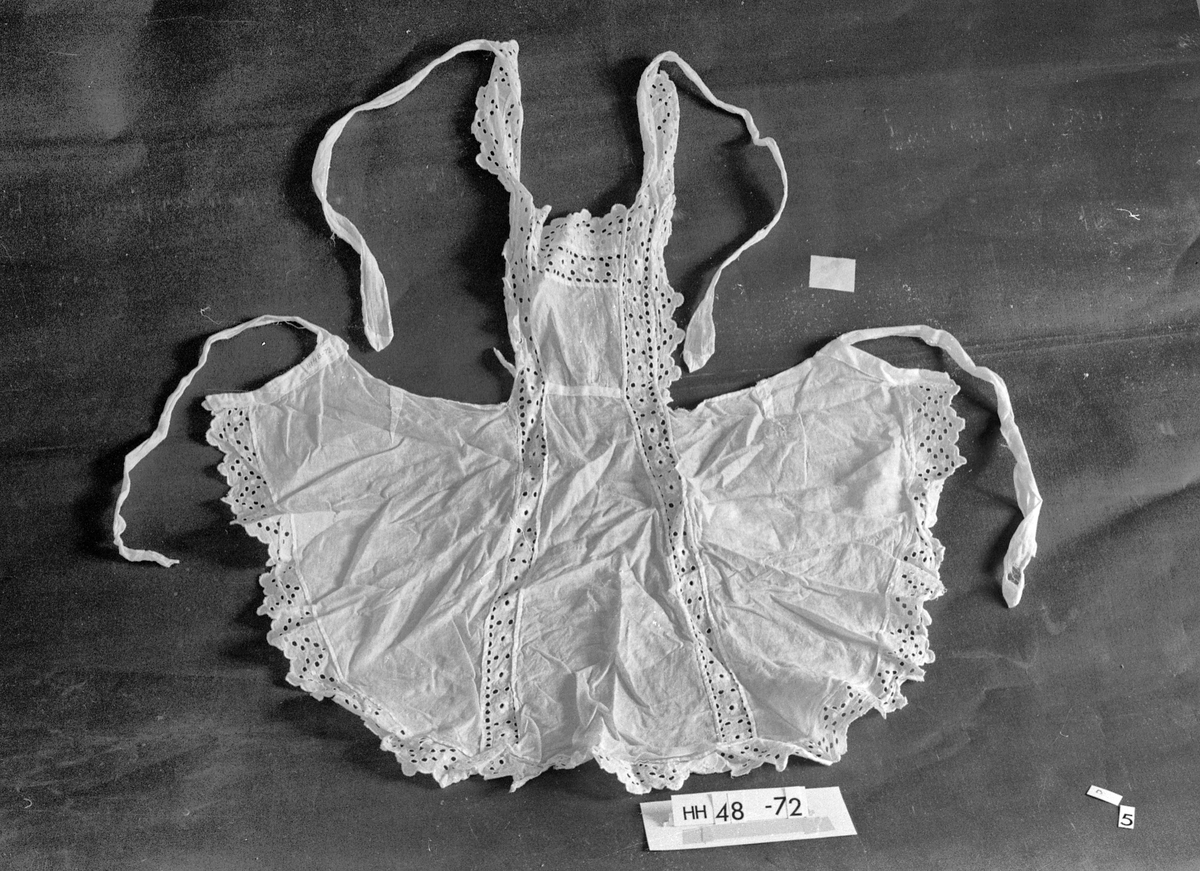 Batist bomull, blonde i sveitsisk broderi.
Blonde: huller, blomster, runger.
Nesten helt kantet med blonde, bredere ved smekken og skuldrene, fortsetter fra smekken som innfelling i forkle.
En lomme.
Skulderbåndene istykker, slitt.
Hvit.
Rustflekker.