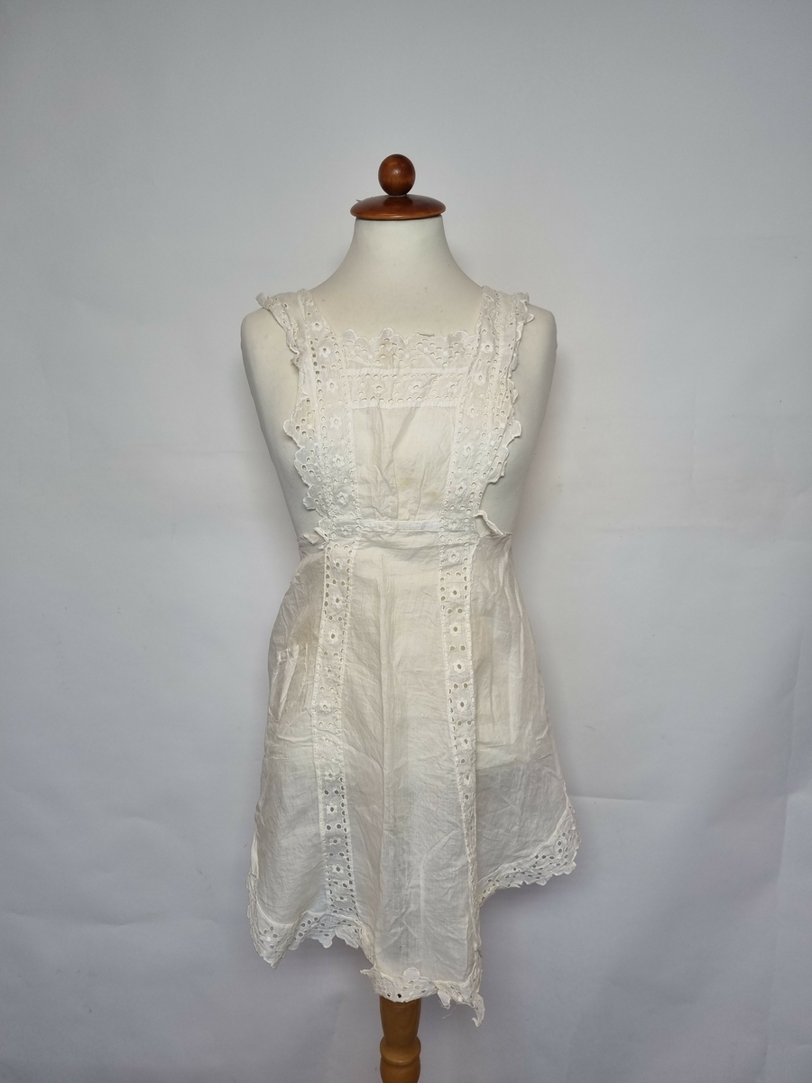Batist bomull, blonde i sveitsisk broderi.
Blonde: huller, blomster, runger.
Nesten helt kantet med blonde, bredere ved smekken og skuldrene, fortsetter fra smekken som innfelling i forkle.
En lomme.
Skulderbåndene istykker, slitt.
Hvit.
Rustflekker.