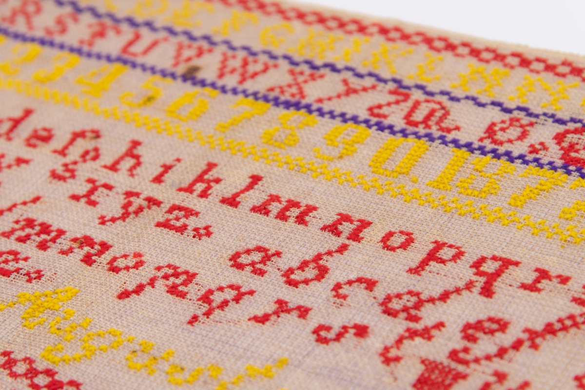 Beige strammei.
Brodert ulltråd, korssting.
Trå rød, gul, sort og lilla.
Brodert: to alfabeter, tall, krone, timeglass. 
"AS" "OS" "Augusta" "1877"