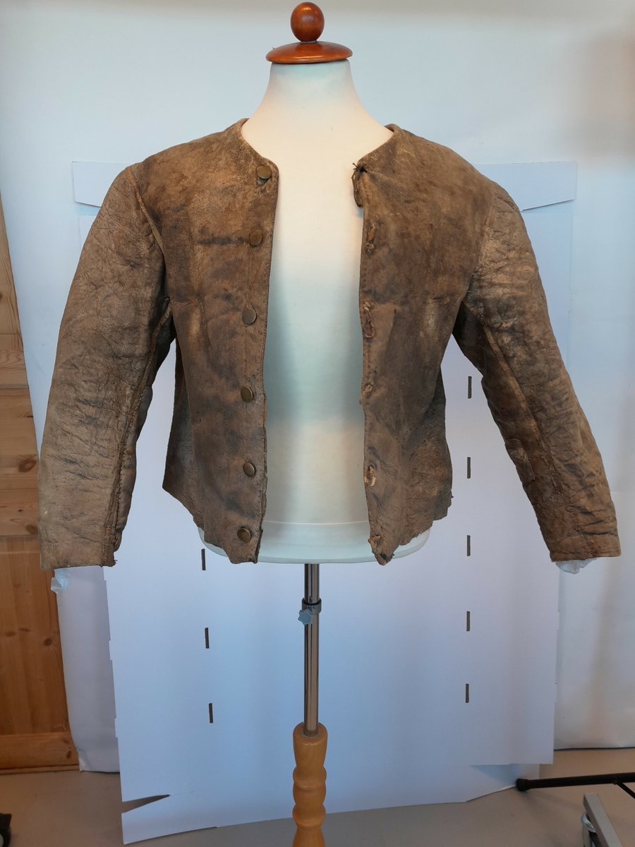 Sauskinn med pelssiden inn.
Håndsydd.
Stykkene er lagt kant i kant og sydd sammen med kastesting med brun lintråd.
Etterpå er alle sømmer skjult av et smalt, en cm bredt skinnbånd, påsydd med lys lintråd, smalere tette kastesting.
Fasongsydde tosømsermer, det høyre med kile.
Halsen er ringet.
Lukningen er midt foran med seks messingknapper og skinnhemper, og har fasongklippet belegg på vrangen. 
Ermene er flere ganger bøtt og reparert.