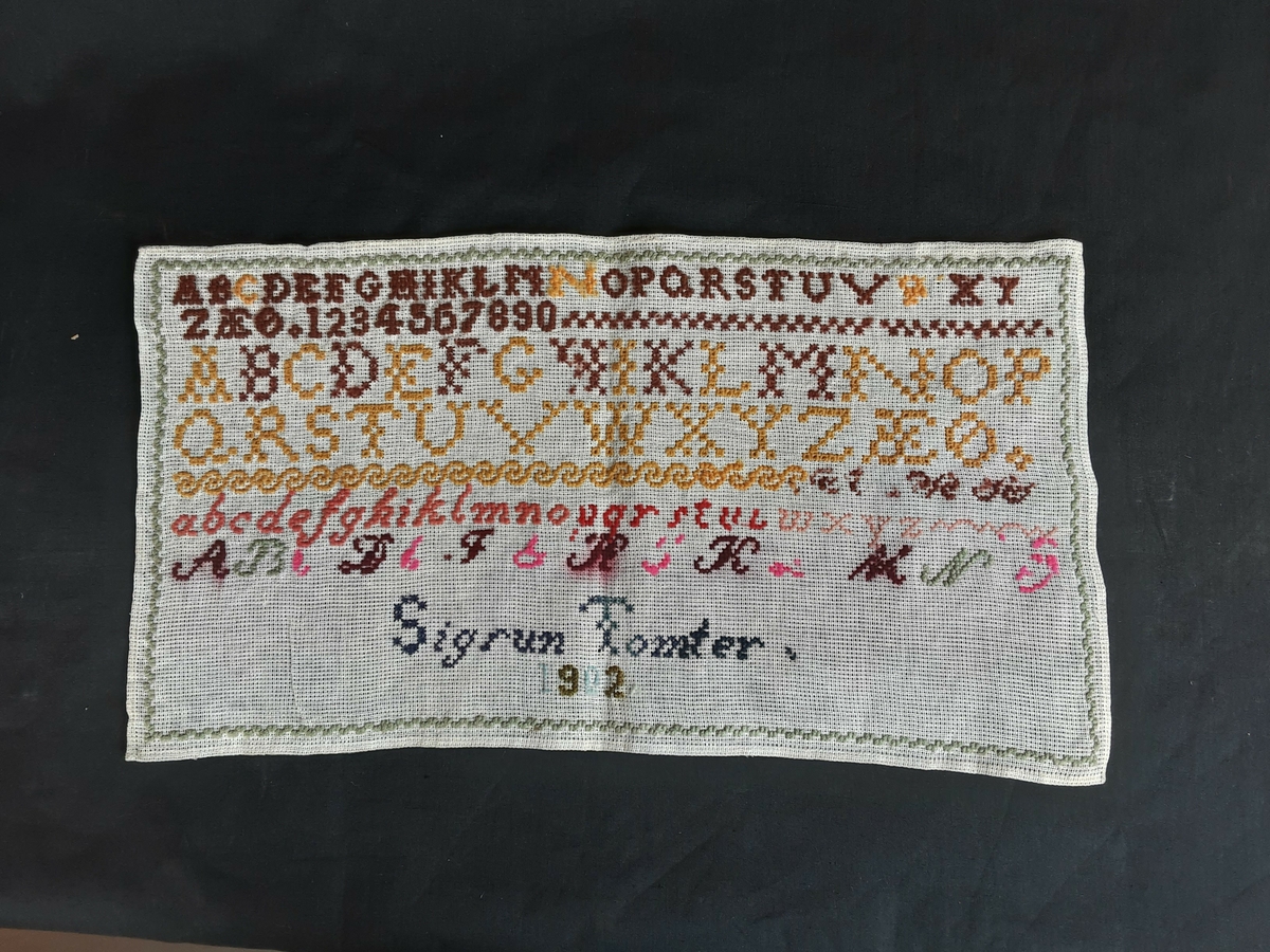 Rektangulært. Brodert alfabet og tall. Store og små bokstaver, samt navn og årstall.