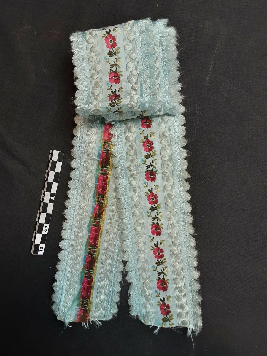 Bånd i lys blå silke med broderier i blomstermotiv. Maskinbroderi. Tungekant