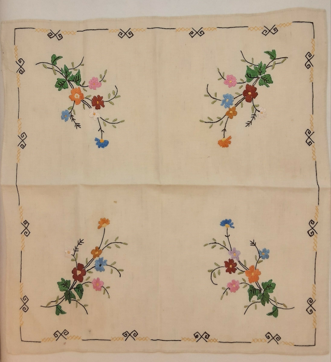 Kvadratisk duk med broderte blomsterbuketter i hvert hjørne.  Broderi rundt i ytterkant i et geometrisk mønster.
