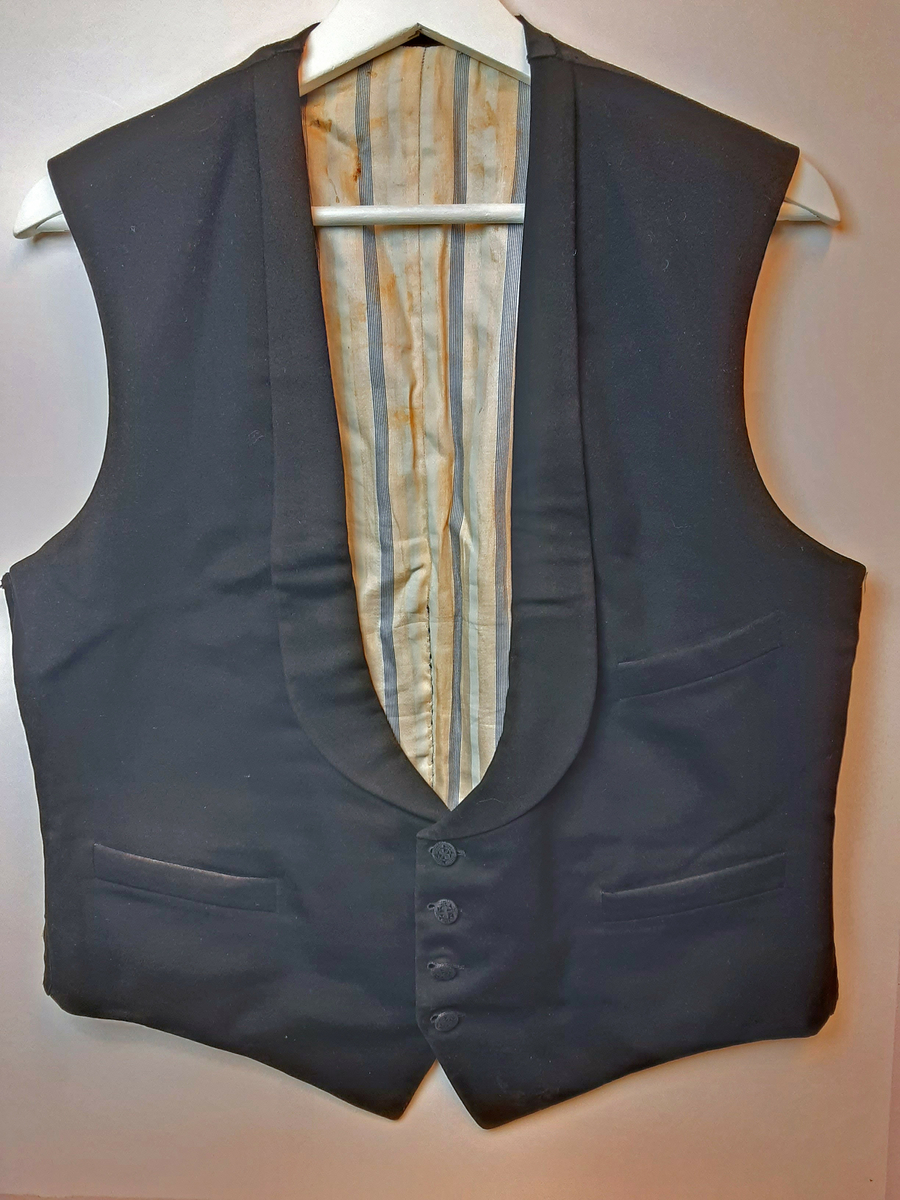 Vest med charleskrage. 3  stikklommer foran. Spensel i ryggen. Knappet foran med 4 knapper trukket med mønstret svart stoff. Hovedstoff framsiden av svart klede; ull, og rygg av silke. Helfóret med stripete bomullsstoff. Splitt bak