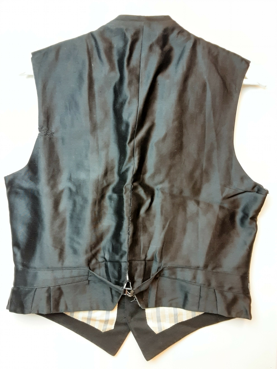 Vest med charleskrage. 3  stikklommer foran. Spensel i ryggen. Knappet foran med 4 knapper trukket med mønstret svart stoff. Hovedstoff framsiden av svart klede; ull, og rygg av silke. Helfóret med stripete bomullsstoff. Splitt bak