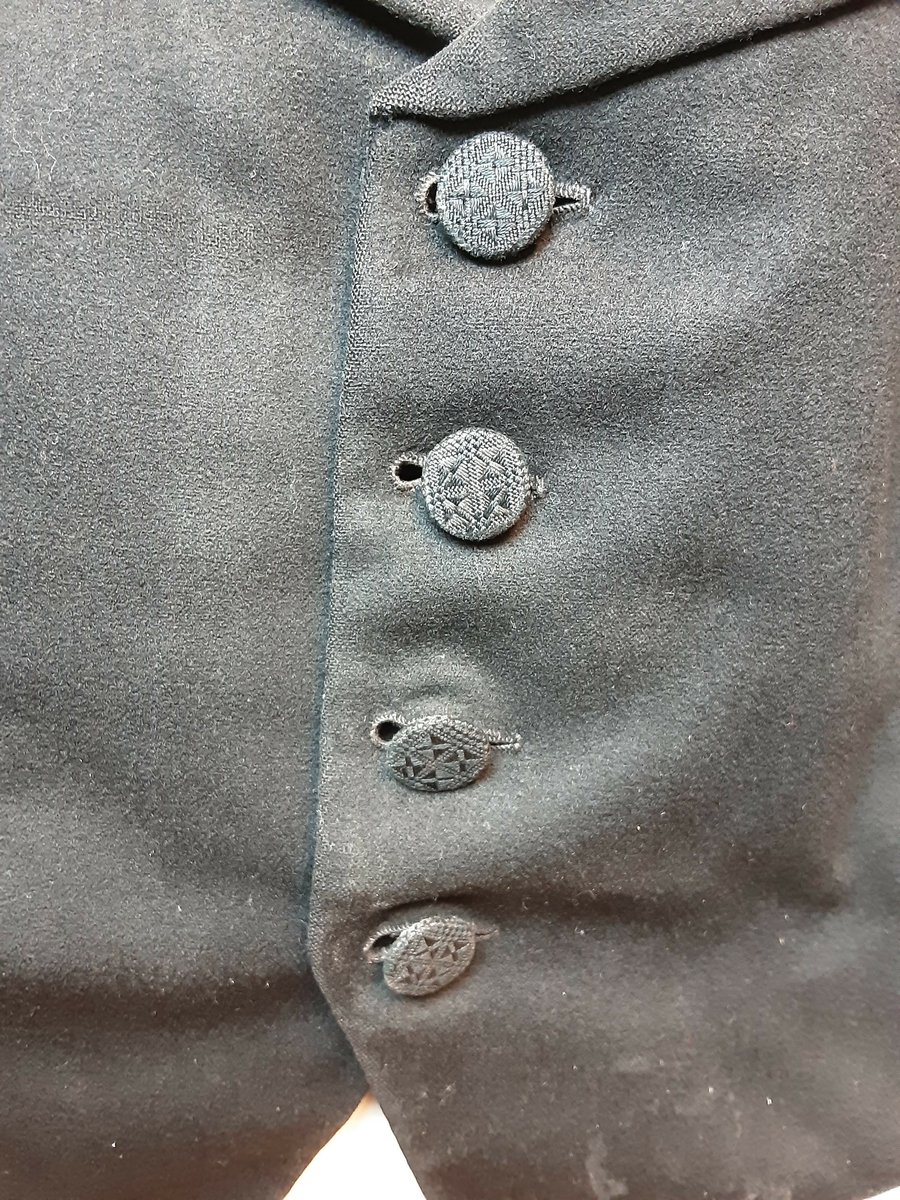 Vest med charleskrage. 3  stikklommer foran. Spensel i ryggen. Knappet foran med 4 knapper trukket med mønstret svart stoff. Hovedstoff framsiden av svart klede; ull, og rygg av silke. Helfóret med stripete bomullsstoff. Splitt bak