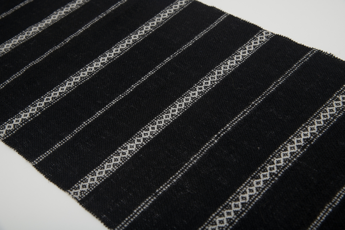 Svart vevd løper med innvevde striper i hvitt; regelmessig annenhver stripe med hvitt mønster og annenhver stripe med 2 hvite innslag.