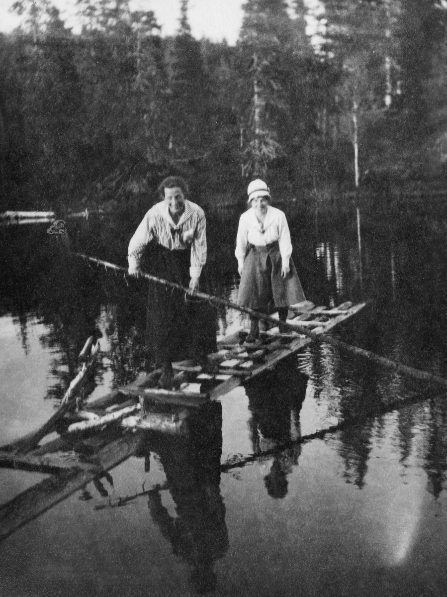 To kvinner på stigeliknende flåte, antakelig på tjern ved Eidet i Grong.  I bakgrunnen granskog. 