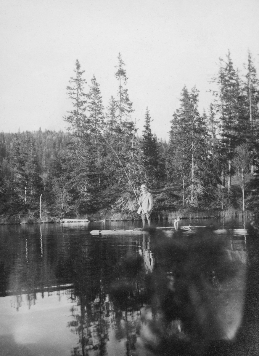 Mannlig sportsfisker med stang.  Mannen står på en flåte på et tjern med granskog i bakgrunnen.  Fotografiet er antakelig tatt på Eidet i Grong. 