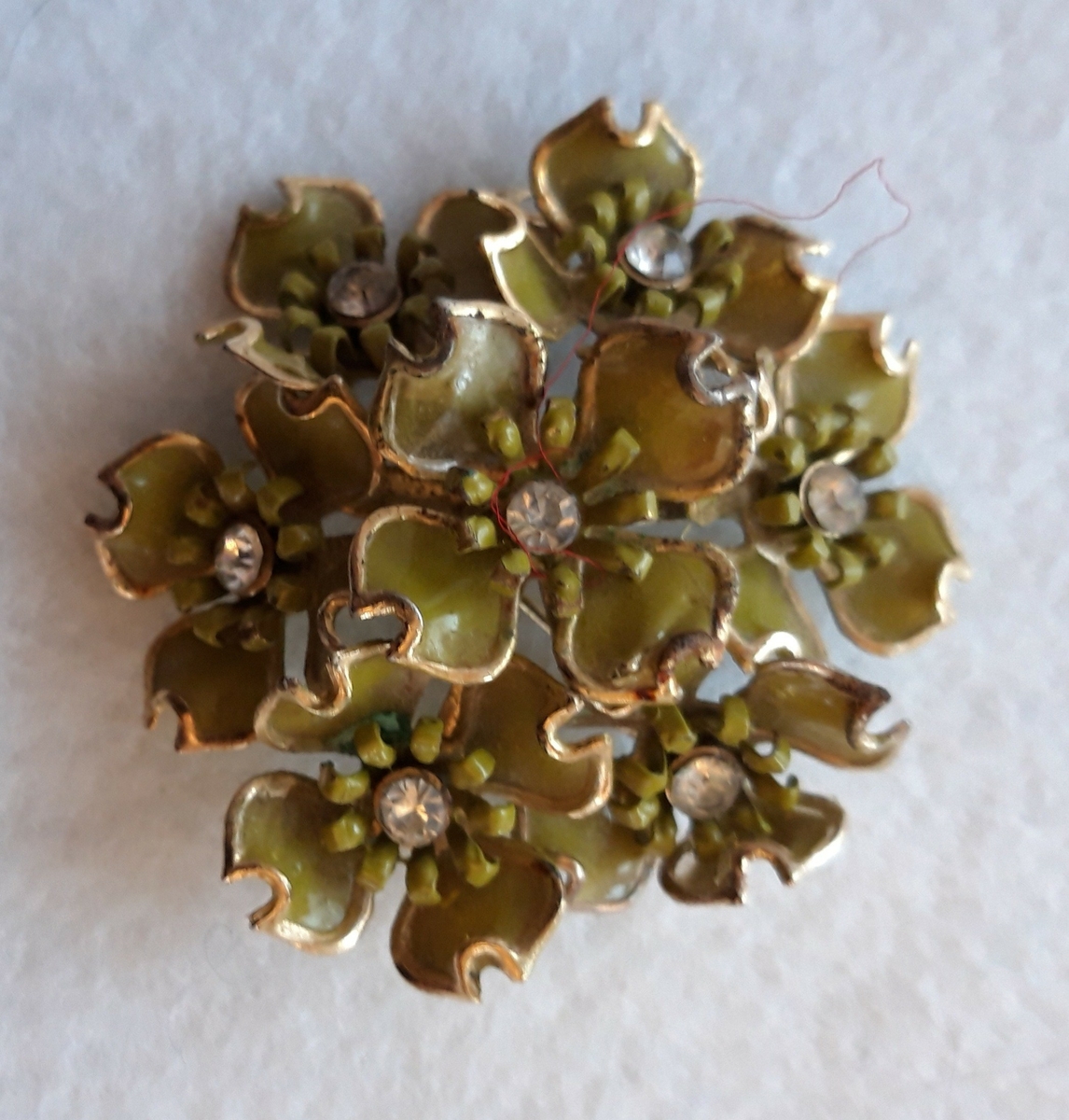 Nål i metall. Sju blomsterkroner er satt sammen og danner en rund nål. Kronbladene er lakkert i en gulgrønn farge. En klar sten er montert i midten av hver blomst.