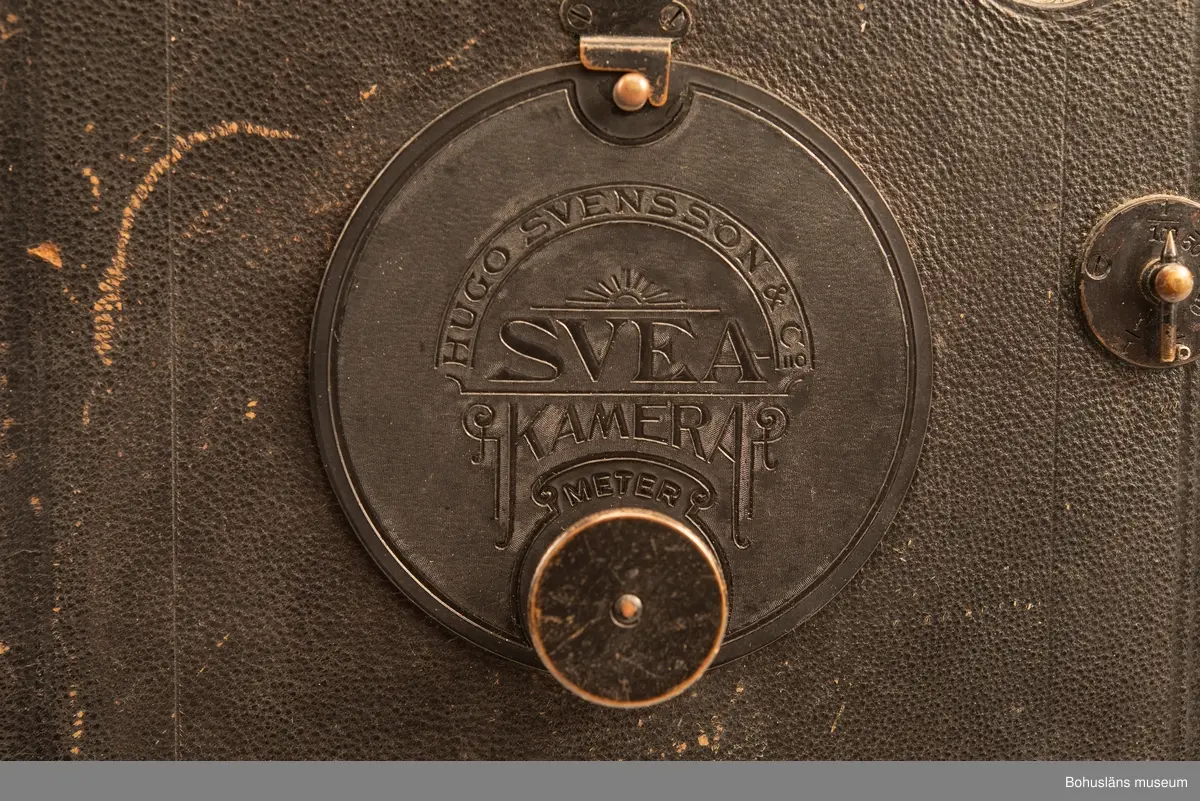 Svart fallplåtskamera av märket SVEA med kamerahus i svart metall. Handtag av läder med inpräglad text: MANUFAKTUR HUGO SVENSSON GÖTENORG
Medföljande originalkartong med klisteretikett: 
HUGO SVENSSON & CO, KAMERAFABRIK, GÖTEBORG
SVEA
NO 14 200