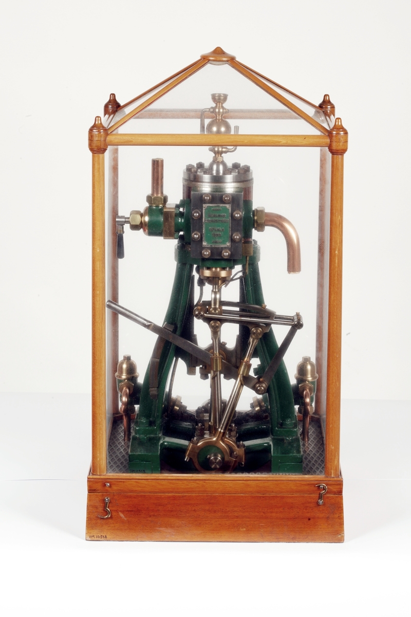 Ångmaskin med grönt järnstativ och övriga delar av stål, mässing och koppar. Skylt på maskinen med text HJALMAR WENNSTRÖM UPSALA 1893. Den står på en fyrkantig bas av polerat trä och är täckt med en rektangulär huv av glas och träribbor fäst med mässingskrokar på basen. Till ångmaskinen hör en "nyckel" (b).